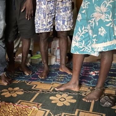 There are four people standing, all of whom have their ankles chained to restrict their movement.