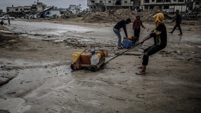 People pull plastic water containers down a muddy street amid destroyed buildings