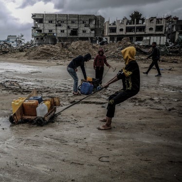 People pull plastic water containers down a muddy street amid destroyed buildings